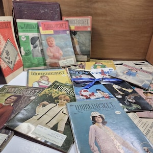May include: A collection of vintage "Workbasket" magazines, featuring colorful covers with illustrations of people, crafts, and seasonal themes. The magazines are stacked and spread out, showcasing various issues from different periods.