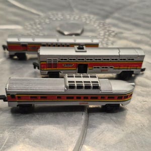 May include: Three toy train cars with silver and red details. The train cars are arranged on a silver surface. The cars have a silver roof and red and yellow accents along the sides.