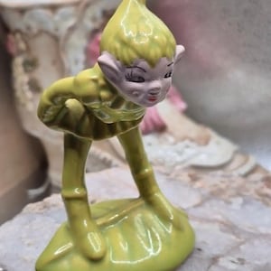 May include: A ceramic figurine of a pointed-ear elf wearing a yellow tunic and hat, standing on a green leaf-shaped base. The elf has a pale face with painted facial features, including dark eyelashes and red lips, and is leaning forward slightly.