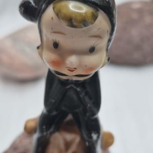 May include: A ceramic figurine of a small character wearing a black hood and black outfit. The character has a pale face with painted black eyes, thin eyebrows, and rosy cheeks. A patch of brown hair is visible under the hood. The figure is seated.