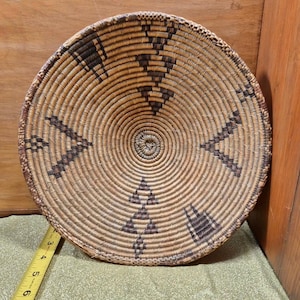 Puede incluir: Cesta redonda tejida con un diseño en espiral. La cesta está hecha de juncos de color natural con patrones geométricos de color marrón oscuro. La cesta tiene aproximadamente 25 cm de diámetro.