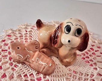 May include: A vintage ceramic salt and pepper shaker set featuring a dog and a shoe. The dog has large black eyes and brown ears. The shoe is light brown. The set is on a pink and white crocheted doily.