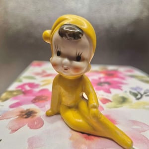 Puede incluir: Una pequeña figura de cerámica de un personaje sentado con un traje amarillo brillante con capucha. La figura tiene un rostro pálido con cabello negro pintado, pestañas largas y mejillas sonrosadas. La capucha tiene un pequeño pompón amarillo.