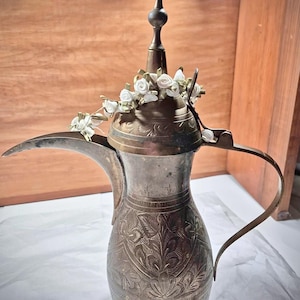 May include: A silver-colored, ornate coffee pot with a long spout and handle. The pot features intricate floral engravings and is adorned with a garland of small white roses around the top. The pot has a tall, pointed finial.