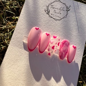 May include: Pink and white oval-shaped press-on nails with a pink heart design. The nails are displayed on a white background with the text "Twinkle Nails by Lynn".