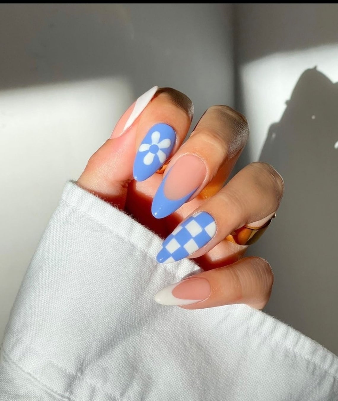 May include: Close-up of a hand with blue and white checkered nail polish on one finger, a white flower design on another finger, and a white tip on another finger.
