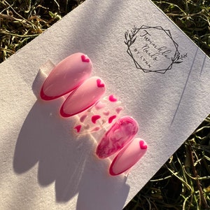 May include: Pink and white press-on nails with a pink heart design. The nails are on a white background with a logo that says "Twinkle Nails by Lynn".