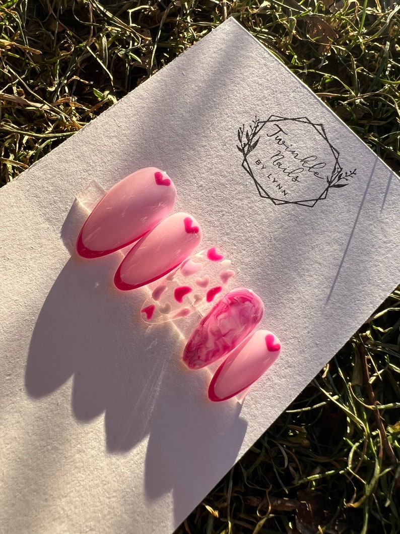 May include: Pink and white press-on nails with a pink heart design. The nails are on a white background with a logo that says "Twinkle Nails by Lynn".
