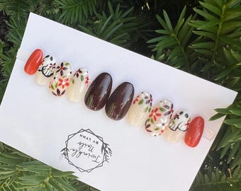 May include: A set of press-on nails arranged on a white card, featuring a variety of designs. Some nails are solid colors, including a vibrant orange and a deep brown. Others showcase floral patterns with red, green, and black accents. The card is labeled "Twinkle Nails by Lynn."