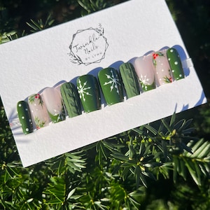 May include: A set of press-on nails with a Christmas theme. The nails feature a variety of designs, including green with white snowflakes, cable knit texture, and polka dots. Some nails have a nude base with holly accents. The brand name "Twinkle Nails by Lynn" is visible.