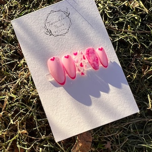May include: Set of five pink press-on nails with a white heart design and a pink glitter accent nail. The nails are displayed on a white card with the text "Twinkle Nails by Lynn".