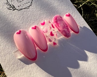 May include: Pink and white heart-shaped nail art with a pink swirl design on one nail. The nail art is displayed on a white card with the text "Twinkle Nails by Lynn".