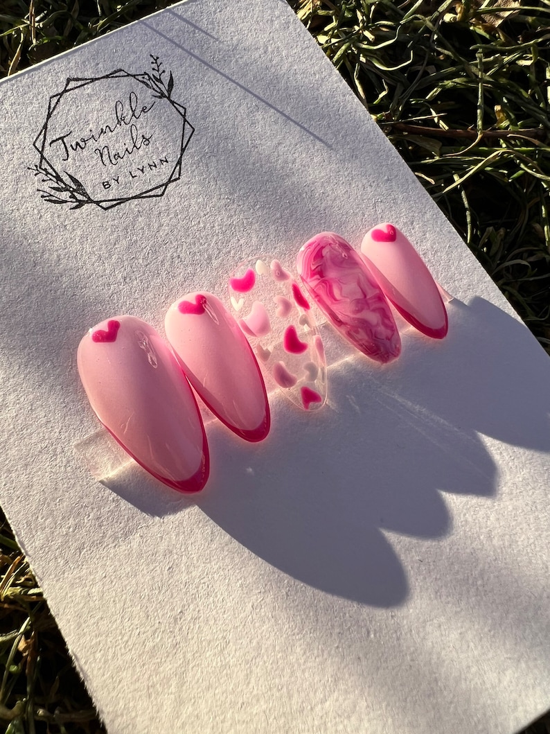 May include: Pink and white heart-shaped nail art with a pink swirl design on one nail. The nail art is displayed on a white card with the text "Twinkle Nails by Lynn".
