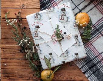 May include: A white gift box wrapped in snowman patterned paper with the text "Let it snow" and a handwritten card that says "Merry Christmas". The box is tied with twine and decorated with a sprig of greenery. The box is sitting on a wooden surface with a plaid blanket and oranges.