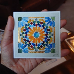May include: A square watercolour painting with a geometric pattern in shades of blue, orange, red, and green, with gold accents. The artwork is held in a hand, showcasing the intricate design and vibrant colours.