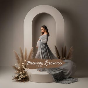 May include: A woman in a gray off-the-shoulder maternity dress stands on a white platform in front of a beige arch. The dress has a long train. Dried grasses and white flowers are arranged around the platform. The text "Maternity Backdrop" is visible.