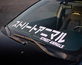 Könnte beinhalten: Nahaufnahme der Windschutzscheibe eines schwarzen Autos mit einem weißen Aufkleber. Der Aufkleber zeigt japanische Schriftzeichen und den englischen Text „STREET ANIMALS“. Der Innenraum des Autos ist sichtbar.