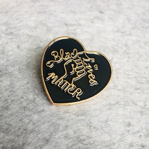 May include: Black Lives Matter pin in the shape of a heart with a raised fist and gold lettering on a black background.