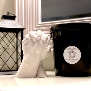 May include: A white plaster hand casting kit with two hands clasped together. The kit is next to a black bucket with a white label that says "Cast Your Life Away" and "Life Casting Studio".