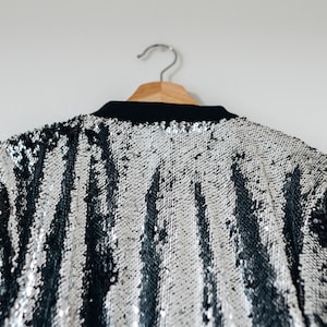 May include: A silver and black sequined jacket with a striped pattern hangs on a wooden hanger.