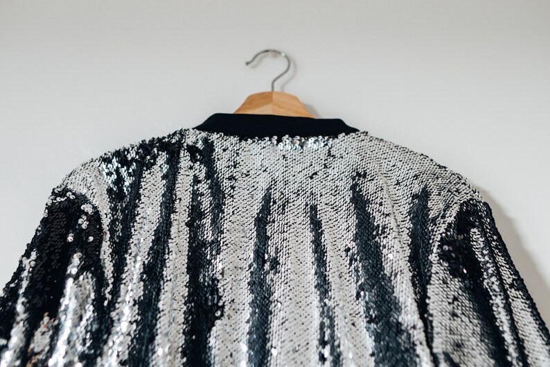 May include: A silver and black sequined jacket with a striped pattern hangs on a wooden hanger.