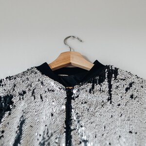 May include: A silver and black sequined bomber jacket hanging on a wooden hanger. The jacket has a zipper closure and a black collar.