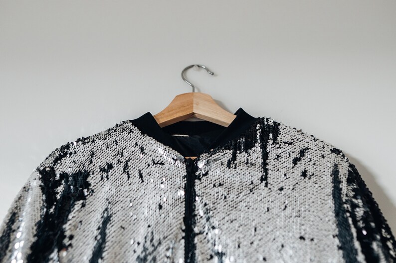 May include: A silver and black sequined bomber jacket hanging on a wooden hanger. The jacket has a zipper closure and a black collar.