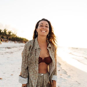 May include: A woman wearing a green floral print shirt and trousers with a brown bikini top, smiling at the camera on a beach.