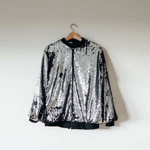 May include: A silver sequined bomber jacket with a black zipper and black lining. The jacket is hanging on a wooden hanger against a white wall.
