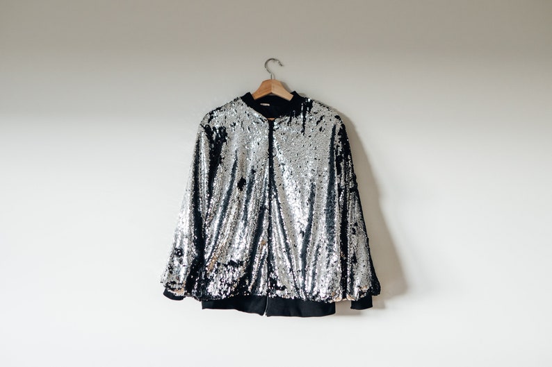 May include: A silver sequined bomber jacket with a black zipper and black lining. The jacket is hanging on a wooden hanger against a white wall.