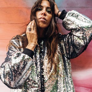 May include: A woman with long brown hair is wearing a silver sequined bomber jacket. The jacket has a black zipper and black trim around the collar and cuffs. The woman is standing in front of a colorful wall.