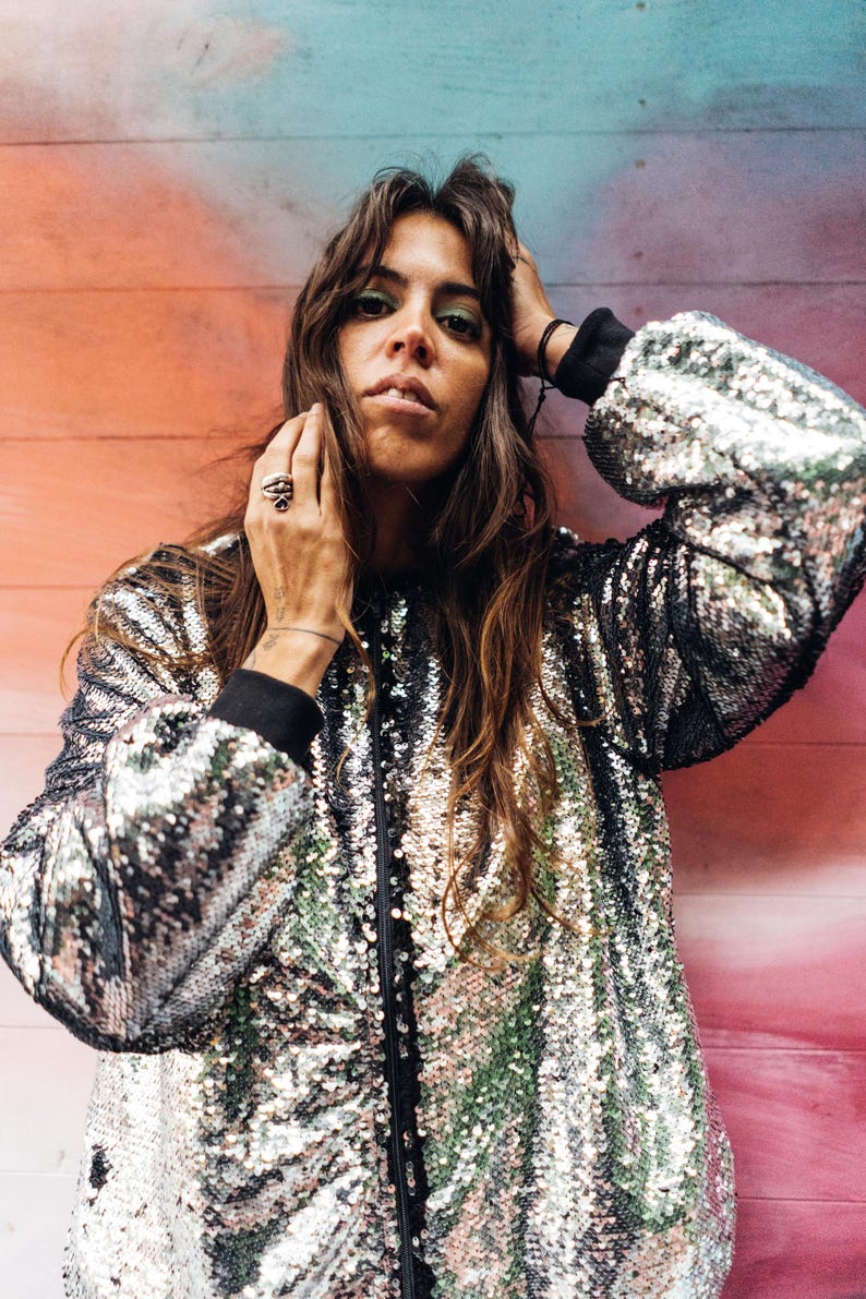 May include: A woman with long brown hair is wearing a silver sequined bomber jacket. The jacket has a black zipper and black trim around the collar and cuffs. The woman is standing in front of a colorful wall.