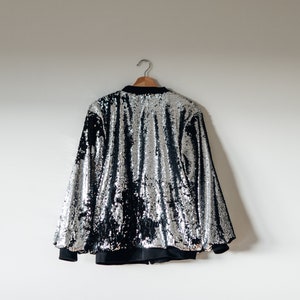May include: A silver sequined bomber jacket with black sleeves and a black zipper. The jacket is hanging on a wooden hanger against a white wall.