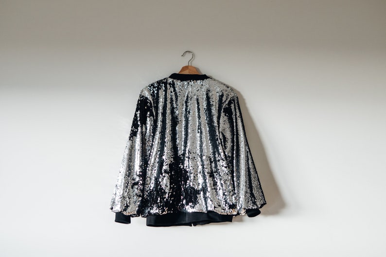 May include: A silver sequined bomber jacket with black sleeves and a black zipper. The jacket is hanging on a wooden hanger against a white wall.