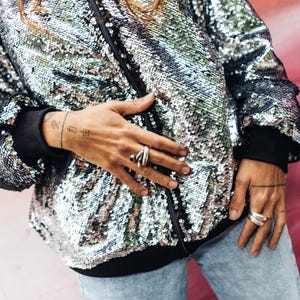 May include: A silver sequined bomber jacket with a black zipper and black trim. The jacket is worn by a person with long brown hair and a tattoo on their wrist.