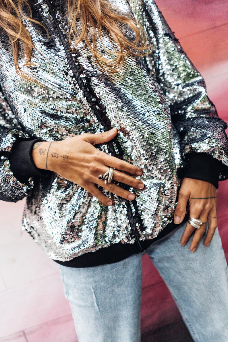 May include: A silver sequined bomber jacket with a black zipper and black trim. The jacket is worn by a person with long brown hair and a tattoo on their wrist.