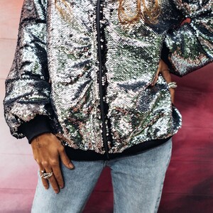 May include: A silver sequined bomber jacket with a zipper closure. The jacket has a black lining and is worn over a pair of blue jeans.