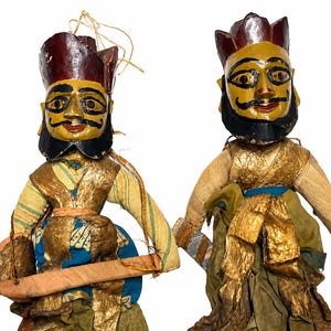 May include: Two vintage hand-painted wooden puppets. Each puppet has a yellow face with black facial hair, a burgundy crown, and is dressed in gold and blue fabric. One puppet holds a wooden instrument.