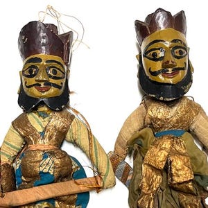 May include: Two vintage hand-carved wooden puppets with painted faces, black beards, and ornate headdresses. The puppets wear gold and blue fabric costumes. The puppets are holding a wooden bar. The puppets are on a white background.