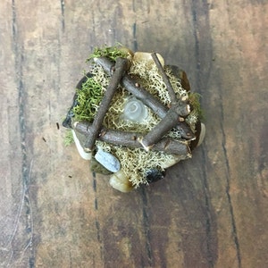May include: A miniature campfire made of small twigs, moss, and stones. The campfire is lit with a small white light in the center.