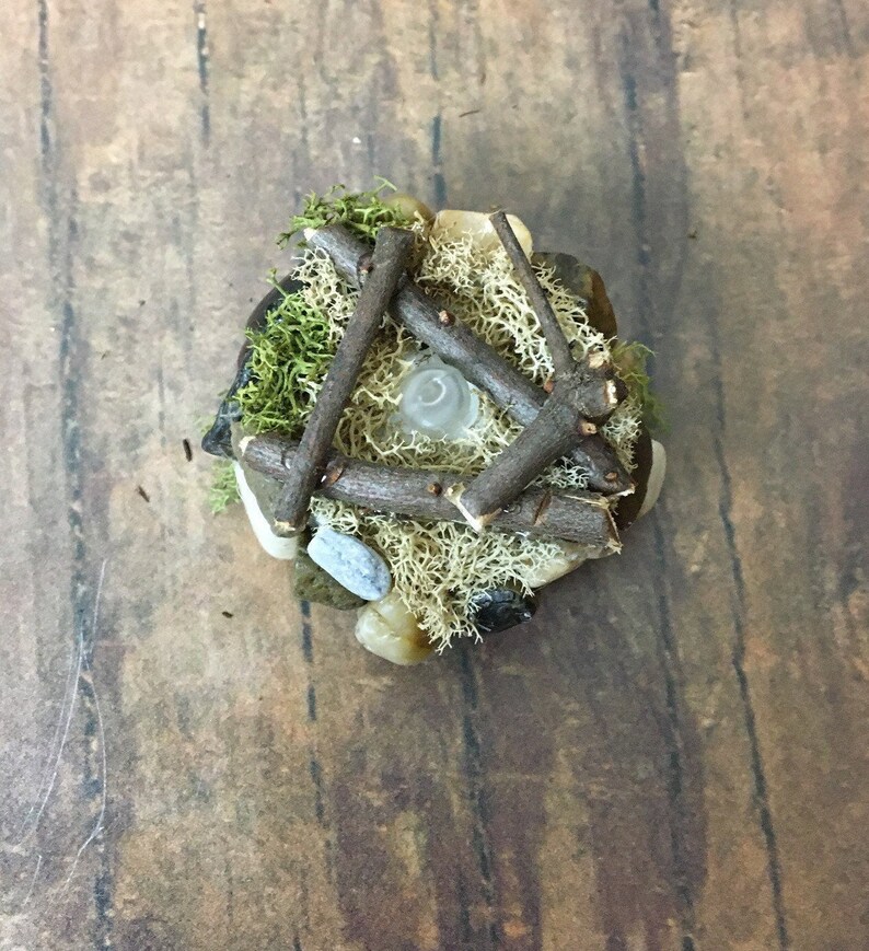 May include: A miniature campfire made of small twigs, moss, and stones. The campfire is lit with a small white light in the center.