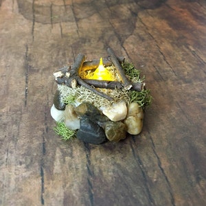 May include: A miniature campfire with a glowing LED light, surrounded by small rocks and moss.