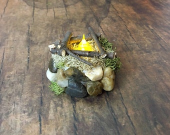 May include: A miniature campfire with a glowing LED light, surrounded by small rocks and moss.
