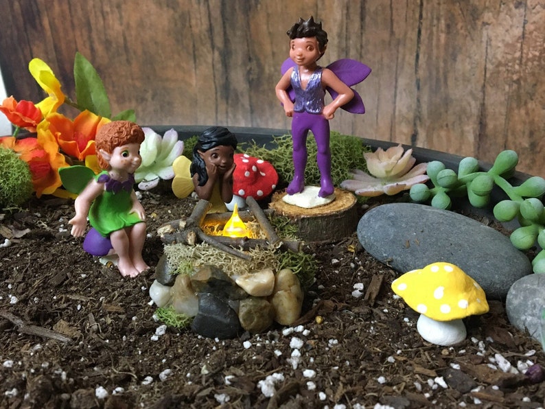 May include: Three fairy figurines in a miniature garden setting. One fairy is purple and has wings, another is green and has wings, and the third is brown and has no wings. The fairies are gathered around a small campfire.