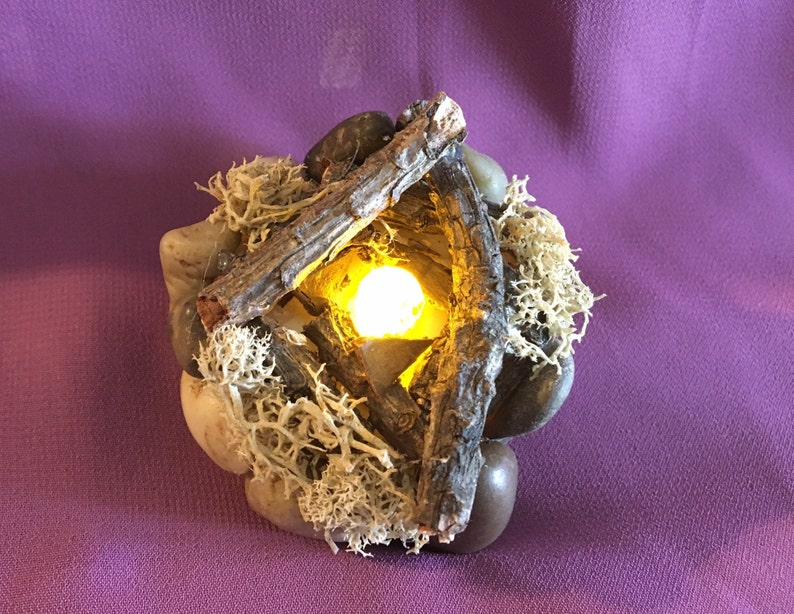 May include: A small, decorative tabletop fire pit made of wood, rocks, and moss. The fire pit has a small, yellow LED light in the center.