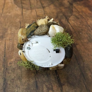 May include: A small, round, white light with a battery compartment on the back. The light is surrounded by small rocks and moss.