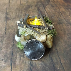 May include: A miniature campfire with a yellow LED light, surrounded by small rocks and moss. A US quarter is included for scale.