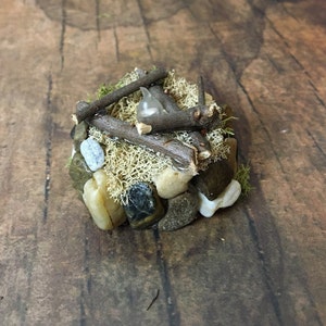 May include: A miniature campfire made of small twigs, rocks, and moss. A small white light bulb is placed in the center of the campfire.