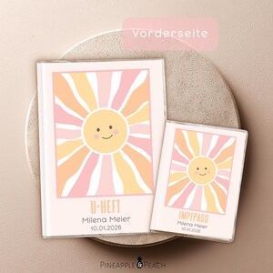 Könnte beinhalten: Zwei rechteckige Schutzhüllen für Dokumente mit einem lächelnden Sonnengrafik-Motiv in Rosa, Orange und Weiß. Die größere Hülle ist mit U-HEFT beschriftet, die kleinere mit IMPFPASS, beide personalisiert mit dem Namen Milena Meier und dem Datum 10.01.2026.