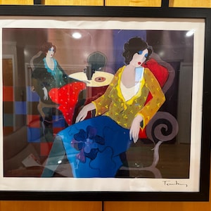 Può includere: Opera d'arte incorniciata con due figure in una stanza. Una figura è seduta su una sedia rossa, con un top giallo e una gonna blu. L'altra figura è sullo sfondo. L'opera d'arte ha uno stile astratto e colorato con una firma in basso a destra.